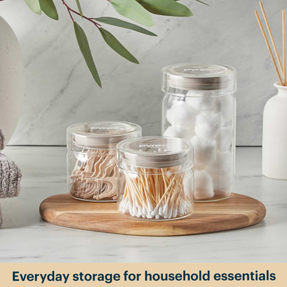 Clear jars with lids containing household essentials on a wooden tray with a neutral background.